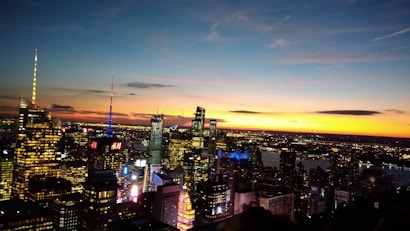 A vibrant cityscape at dusk featuring numerous illuminated skyscrapers and billboards. The skyline is set against a dramatic sunset with a blend of deep oranges, blues, and hints of pink, casting a warm glow over the urban landscape and reflecting on a nearby body of water.