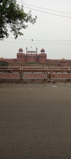 A large historic fort with red sandstone architecture in the background, situated beyond barriers displaying 'Delhi Police'. The fort features multiple domes and an Indian flag flying at the top. Trees partially frame the image in the upper left corner, and a bird is perched on a wire.