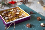 An open box of assorted chocolates is placed on a textured green mat with some candies scattered around. The chocolates are in various shapes and designs, featuring a mix of dark, milk, and white chocolate. The ambiance is cozy with small festive decorations, such as a red star and pine cones, adding a holiday feel. There are also fairy lights and a rustic wooden surface in the background.