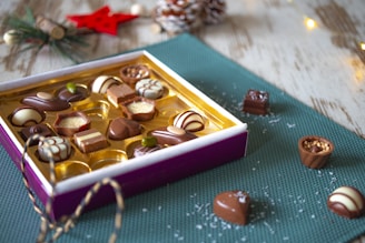 An open box of assorted chocolates is placed on a textured green mat with some candies scattered around. The chocolates are in various shapes and designs, featuring a mix of dark, milk, and white chocolate. The ambiance is cozy with small festive decorations, such as a red star and pine cones, adding a holiday feel. There are also fairy lights and a rustic wooden surface in the background.