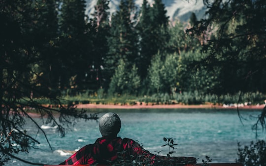 person in red and black jacket sitting on black bench near body of water during daytime in Town Of Banff Canada