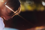 A young child with a serene and introspective expression is captured in profile, illuminated by a warm, golden light. The sun's rays create striking highlights on the hair, casting gentle shadows on the face. The background is softly blurred with hints of greenery, enhancing the focus on the child.
