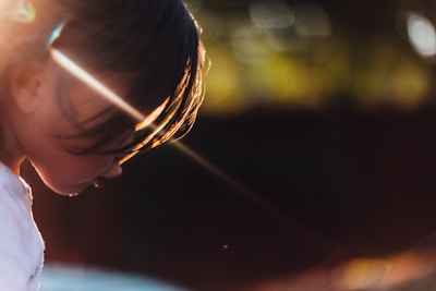 A young child with a serene and introspective expression is captured in profile, illuminated by a warm, golden light. The sun's rays create striking highlights on the hair, casting gentle shadows on the face. The background is softly blurred with hints of greenery, enhancing the focus on the child.