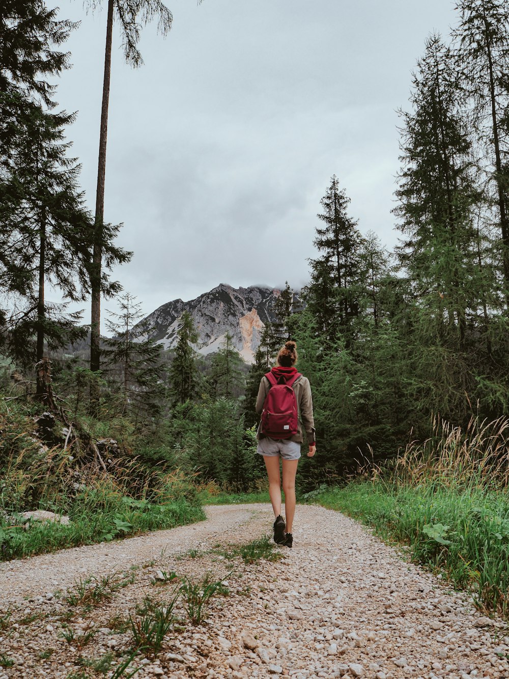 woman in red jacket and brown shorts standing on pathway near green trees and mountain during