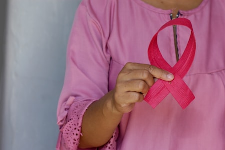 A person wearing a pink shirt holds a pink ribbon, often associated with breast cancer awareness, between their fingers. The focus is on the hand and ribbon, highlighting the texture and color of the fabric.