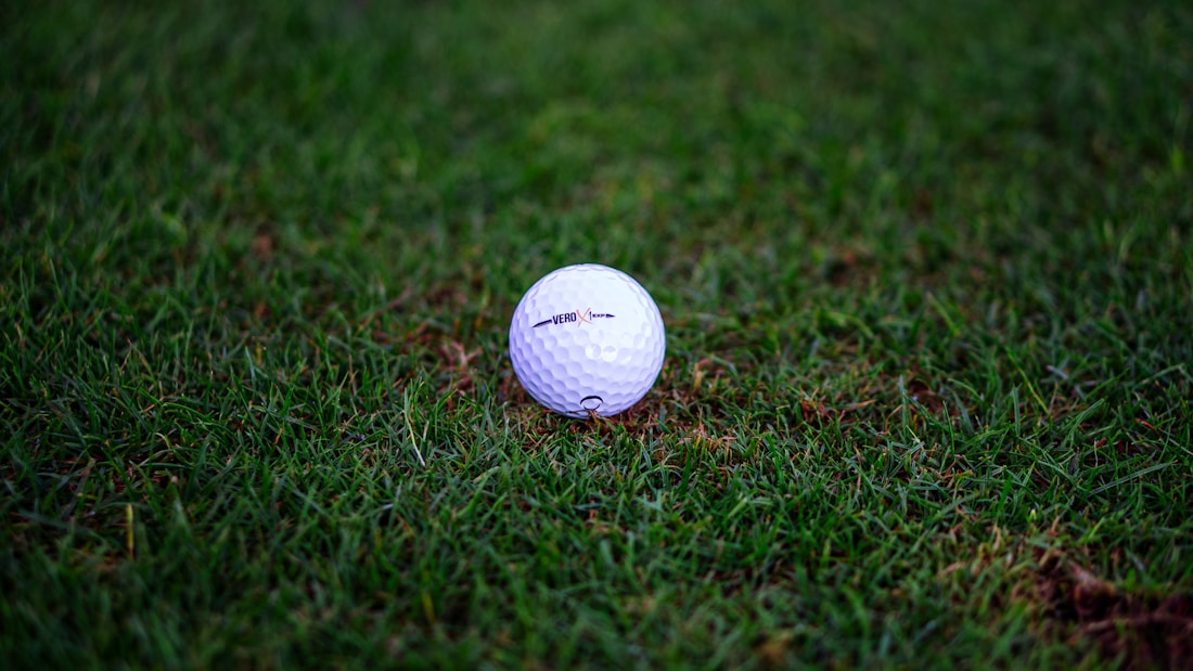 A white golf ball resting on a lush green lawn with a slightly worn surface. The grass appears freshly cut, and the golf ball features a textured, dimpled surface.