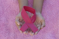 Hands are gently holding a pink ribbon, symbolizing awareness, on a textured pink fabric background.