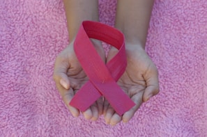 Hands are gently holding a pink ribbon, symbolizing awareness, on a textured pink fabric background.