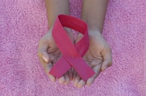Hands are gently holding a pink ribbon, symbolizing awareness, on a textured pink fabric background.