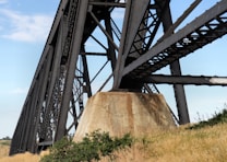 A large, robust steel bridge with intricate lattice design spans across a clear blue sky. The bridge is supported by a substantial concrete pillar which anchors it to the ground. Surrounding the base are patches of green shrubs and dry grasses, giving an impression of a rural setting.