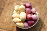 A clear plastic container holds peeled cloves of garlic alongside small, peeled red onions. The arrangement is set against a textured wooden surface, providing a warm and rustic feel.