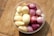 A clear plastic container holds peeled cloves of garlic alongside small, peeled red onions. The arrangement is set against a textured wooden surface, providing a warm and rustic feel.