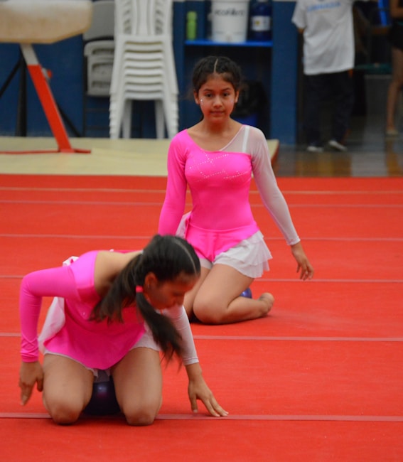 Two young girls in bright pink leotards are performing on a red gymnastics floor. One girl is kneeling and holding a ball, while the other is sitting upright, both wearing white skirts attached to their leotards. In the background, there are stacked chairs and some gymnasium equipment.
