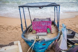 A rustic wooden boat with a colorful sign displaying painted words and images rests on a sandy beach near the shoreline. The boat has a simple canopy and appears weathered, with peeling paint in shades of blue and purple. Ocean waves gently approach the shore in the background.