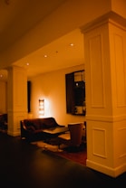 A dimly lit lounge area features plush seating and a central coffee table. The warm lighting creates a cozy atmosphere with a lamp casting a soft glow on the wall. A decorative mirror and soft furniture enhance the inviting ambiance.