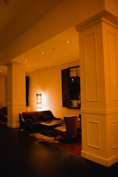A dimly lit lounge area features plush seating and a central coffee table. The warm lighting creates a cozy atmosphere with a lamp casting a soft glow on the wall. A decorative mirror and soft furniture enhance the inviting ambiance.