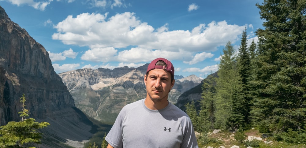 homme en t-shirt à col rond blanc debout près des arbres verts et des montagnes pendant la journée