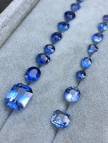 A collection of blue gemstones arranged in two rows on a soft gray surface. The gemstones vary in size and shape, including both round and rectangular cuts, with a brilliant shine.