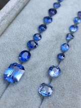 A collection of blue gemstones arranged in two rows on a soft gray surface. The gemstones vary in size and shape, including both round and rectangular cuts, with a brilliant shine.