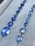 A collection of blue gemstones arranged in two rows on a soft gray surface. The gemstones vary in size and shape, including both round and rectangular cuts, with a brilliant shine.