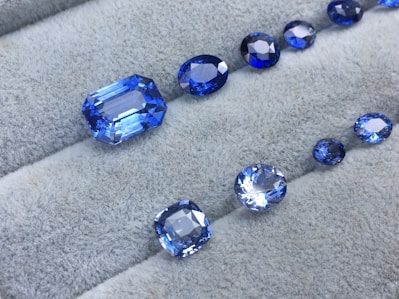A collection of blue gemstones in various shapes and sizes is displayed on a light gray, textured surface. The gems exist in a range of cuts, including oval and rectangular, showcasing their reflective and faceted surfaces that shimmer in different shades of blue.