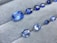 A collection of blue gemstones in various shapes and sizes is displayed on a light gray, textured surface. The gems exist in a range of cuts, including oval and rectangular, showcasing their reflective and faceted surfaces that shimmer in different shades of blue.