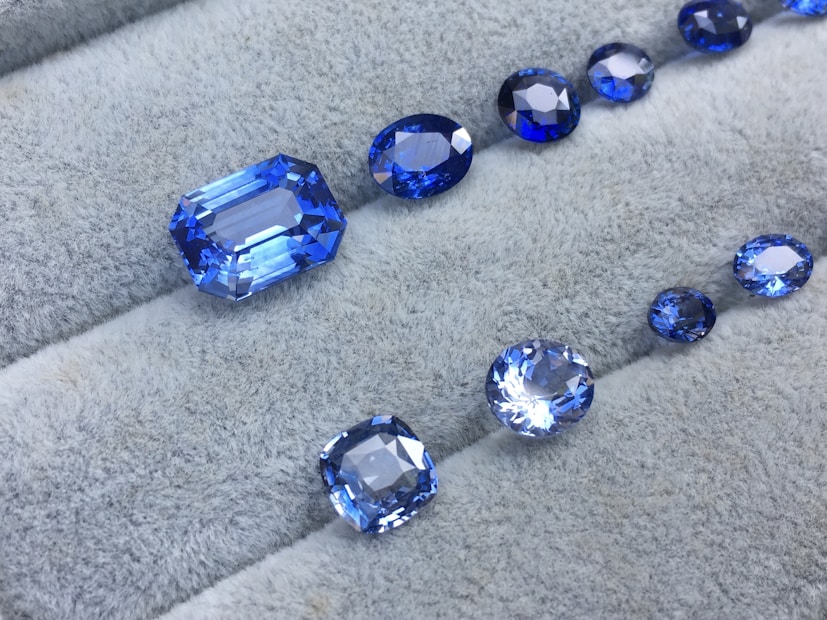 A collection of blue gemstones in various shapes and sizes is displayed on a light gray, textured surface. The gems exist in a range of cuts, including oval and rectangular, showcasing their reflective and faceted surfaces that shimmer in different shades of blue.