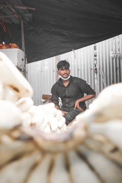 A person sits under a dark canopy beside a stack of bananas, with some oranges visible in the background. The setting appears to be an open market, and a corrugated metal wall is behind him. The person is dressed in a dark shirt and wears a mask positioned under the chin.