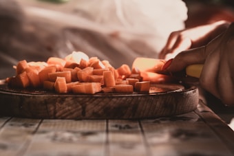 A close-up of hands cutting orange vegetables, likely carrots, on a round wooden cutting board. The scene is warmly lit, highlighting the texture and color of the chopped pieces.