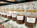 A row of glass jars filled with different types of coffee beans, each labeled with a specific blend, such as Viennese Blend, Oriental Espresso Blend, and Arabica Plantation-AAA. The jars are neatly aligned on a glass shelf, and the coffee beans inside are varying shades of brown.