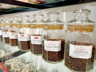A row of glass jars filled with different types of coffee beans, each labeled with a specific blend, such as Viennese Blend, Oriental Espresso Blend, and Arabica Plantation-AAA. The jars are neatly aligned on a glass shelf, and the coffee beans inside are varying shades of brown.