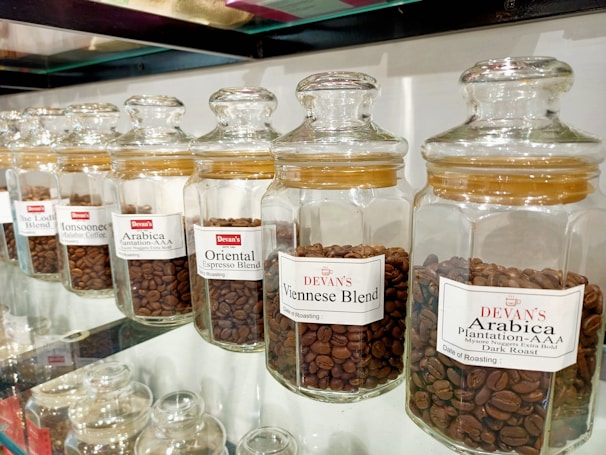 A row of glass jars filled with different types of coffee beans, each labeled with a specific blend, such as Viennese Blend, Oriental Espresso Blend, and Arabica Plantation-AAA. The jars are neatly aligned on a glass shelf, and the coffee beans inside are varying shades of brown.