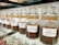 A row of glass jars filled with different types of coffee beans, each labeled with a specific blend, such as Viennese Blend, Oriental Espresso Blend, and Arabica Plantation-AAA. The jars are neatly aligned on a glass shelf, and the coffee beans inside are varying shades of brown.