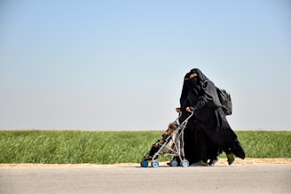 A woman dressed in a black niqab and abaya is pushing a stroller with a child seated in it. The setting is outdoors, alongside a road bordered by green grass under a clear blue sky. The woman carries a backpack, and the scene conveys movement.