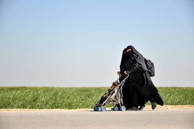 A woman dressed in a black niqab and abaya is pushing a stroller with a child seated in it. The setting is outdoors, alongside a road bordered by green grass under a clear blue sky. The woman carries a backpack, and the scene conveys movement.