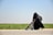 A woman dressed in a black niqab and abaya is pushing a stroller with a child seated in it. The setting is outdoors, alongside a road bordered by green grass under a clear blue sky. The woman carries a backpack, and the scene conveys movement.
