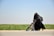 A woman dressed in a black niqab and abaya is pushing a stroller with a child seated in it. The setting is outdoors, alongside a road bordered by green grass under a clear blue sky. The woman carries a backpack, and the scene conveys movement.