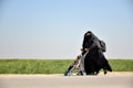 A woman dressed in a black niqab and abaya is pushing a stroller with a child seated in it. The setting is outdoors, alongside a road bordered by green grass under a clear blue sky. The woman carries a backpack, and the scene conveys movement.