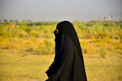 A person wearing a full black garment, possibly a hijab or abaya, walking outdoors in a grassy field with a blurred background featuring green and yellow foliage under a clear sky.