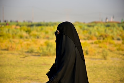 A person wearing a full black garment, possibly a hijab or abaya, walking outdoors in a grassy field with a blurred background featuring green and yellow foliage under a clear sky.