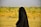 A person wearing a full black garment, possibly a hijab or abaya, walking outdoors in a grassy field with a blurred background featuring green and yellow foliage under a clear sky.