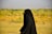 A person wearing a full black garment, possibly a hijab or abaya, walking outdoors in a grassy field with a blurred background featuring green and yellow foliage under a clear sky.