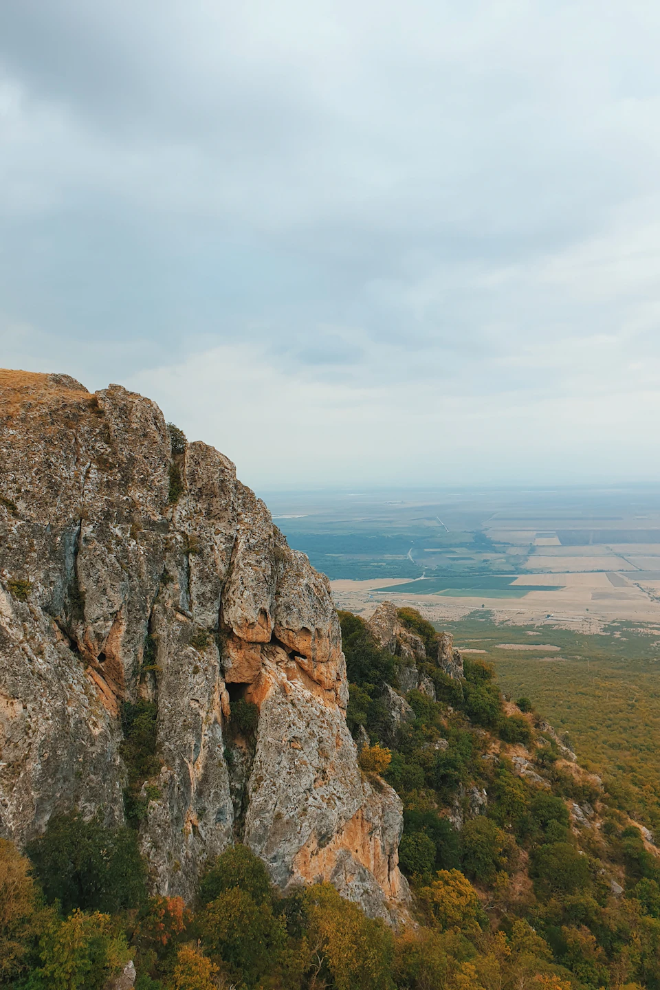 The image depicts a rocky cliff face with patches of green vegetation, set against an overcast sky and a valley below. The perspective is from a high vantage point, providing a sweeping view of the landscape.