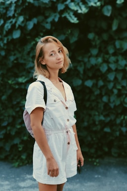 A person with short blonde hair is standing outdoors against a backdrop of lush green leaves. They are wearing a white short-sleeved romper with button details and have a backpack on one shoulder.