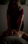 A person is giving a massage in a dimly lit room. The focus is on the hands applying pressure on the back of the individual lying down. The atmosphere appears calm and therapeutic.