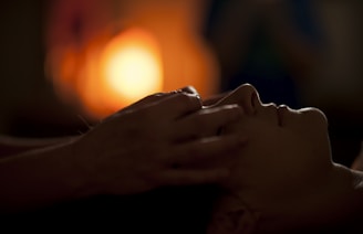 A person lies down with eyes closed, while another person places hands gently on their forehead. The background is softly lit with a warm, orange glow, creating a calming atmosphere.