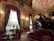 A lavish, opulent room adorned with ornate gold decorations and intricate moldings. Rich, deep-red velvet curtains drape over large windows, while numerous crystal chandeliers hang from the ceiling, casting a warm glow. The room features elaborate furniture, including carved wooden chairs upholstered in matching red velvet, small round tables, and a large plush sofa. Luxurious decor includes large vases and indoor plants that add a touch of greenery.