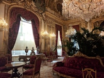 A lavish, opulent room adorned with ornate gold decorations and intricate moldings. Rich, deep-red velvet curtains drape over large windows, while numerous crystal chandeliers hang from the ceiling, casting a warm glow. The room features elaborate furniture, including carved wooden chairs upholstered in matching red velvet, small round tables, and a large plush sofa. Luxurious decor includes large vases and indoor plants that add a touch of greenery.