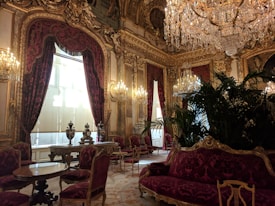 A lavish, opulent room adorned with ornate gold decorations and intricate moldings. Rich, deep-red velvet curtains drape over large windows, while numerous crystal chandeliers hang from the ceiling, casting a warm glow. The room features elaborate furniture, including carved wooden chairs upholstered in matching red velvet, small round tables, and a large plush sofa. Luxurious decor includes large vases and indoor plants that add a touch of greenery.