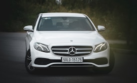 A sleek white Mercedes-Benz car is parked on a dark road with greenery in the background. The car's front view is captured, showcasing its elegant grille and shiny headlights. The license plate reads '50LD-151.75'. The overall setting is dimly lit, highlighting the vehicle's features.